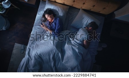 Top View: Young Woman Uses Smartphone in Bed at Night When Her Male Partner Trying to Fall Asleep Beside. Couple Fight, Argue. Addictive World of Social Media, Doom Scrolling, Fake News. Royalty-Free Stock Photo #2229884741