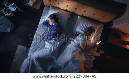 Top View Bedroom Apartment: Man Uses Smartphone in Bed at Night When His Female Partner Trying to Fall Asleep. Couple After Fight, Argument. Addictive World of Social Media, Doom Scrolling, Fake News. Royalty-Free Stock Photo #2229884745