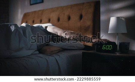 Man Wakes Up, Turns off Alarm Clock with Frustration, after Sleepless Insomniac Night. Early Rising Stressed Man Ready to Face Day of Problem Solving. Focus on the Clock Showing Six A.M. Royalty-Free Stock Photo #2229885559