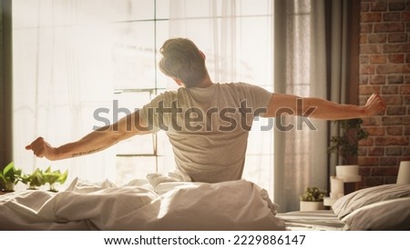 Handsome Young Man Waking up in the Morning, Stretches and Gets Out of Bed, Sun Shines From the Apartment Window in Bedroom, He is Ready for Business Opportunities, Achievements, Adventures Royalty-Free Stock Photo #2229886147