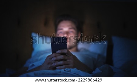 Caucasian Man Uses Smartphone in Bed at Home at Night. Handsome Guy Browsing Social Media, Reading News, Doing Online Shopping, Chatting with Friends Late at Night. Focus on Hand Holding Mobile Phone Royalty-Free Stock Photo #2229886429