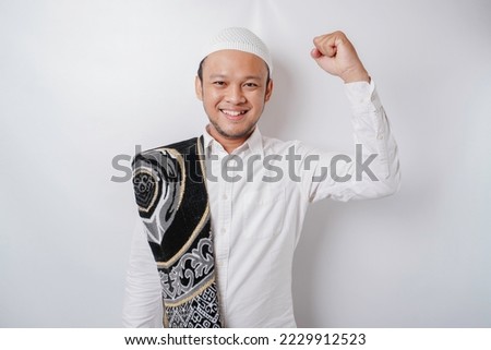 A happy young Asian Muslim man with a prayer rug on his shoulder showing a successful expression isolated by white background