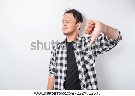 Disappointed Asian man wearing tartan short gives thumbs down hand gesture of approval, isolated by white background