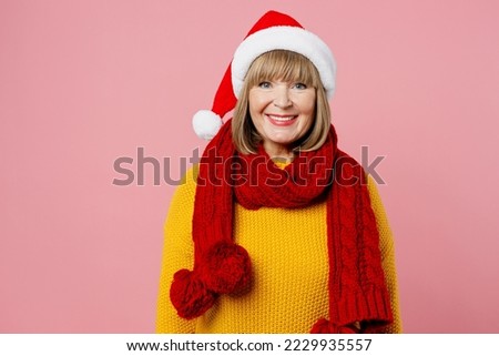 Merry smiling fun positive elderly woman 50s years old wearing yellow knitted sweater red scarf Santa hat posing look camera isolated on plain pink background. Happy New Year Christmas 2023 concept