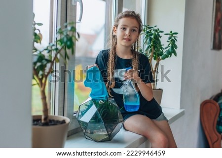 The child is holding cleaning supplies. A teenager helps with household chores. A cute and cheerful girl cleans up with detergents at home. The girl is sitting on the windowsill.
