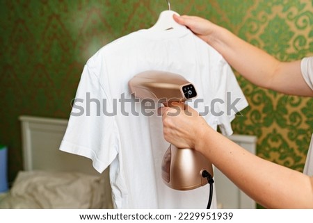 woman irons white t-shirt by portable hand steamer for clothes. household appliances for the home. Royalty-Free Stock Photo #2229951939