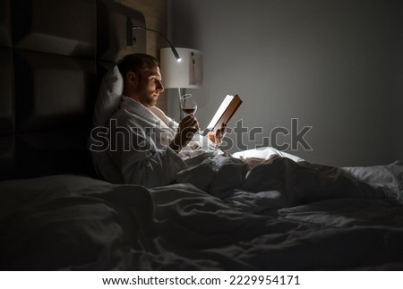 Midle-aged man relaxing rin bed reading book holding a glass of red wine with bedside lamp turned on. Evening relaxation, hobbies, free time concept. Adulthood concept. Royalty-Free Stock Photo #2229954171