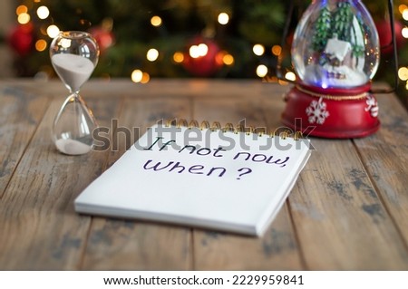 Hourglass and notepad with motivational inscription - if not now, when on the background of Christmas tree. Striving for change, push for development concept