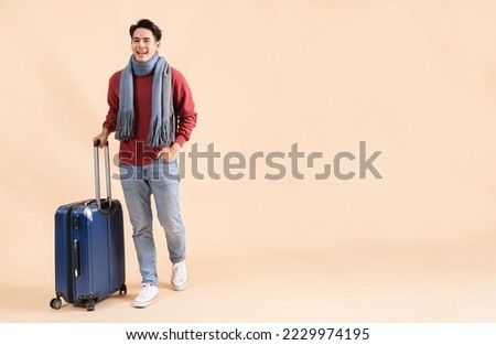 Happy Asian male tourist with luggage in red sweater and scarf enjoying winter vacation isolated on beige color background. Excited fashionable 20s guy going on a trip - Full body length studio shot.