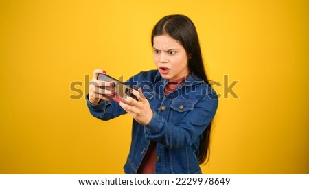 Sad or Angry modern gamer girl using mobile phone, Aggressive or shocked young woman playing video game on smartphone, lost the game, standing isolated over yellow background