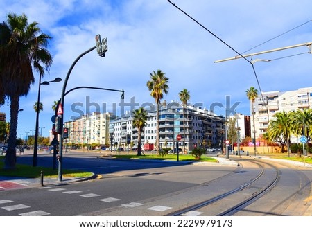 Traffic light over road and pedestrian crossing in city. Tram line and multicolored facades of residential buildings in town among palm trees. Valencia city street, road traffic, cars on road, people.