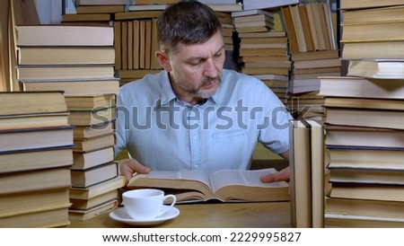 A bearded man reads a book in the library. Stacks of books and cup of tea on table. Concept of Reading and Education.