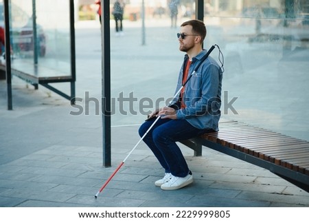 Visually impaired man with walking stick, sitting on bench in city park. Copy space. Royalty-Free Stock Photo #2229999805