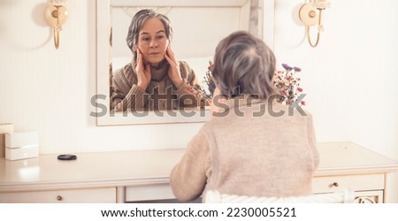 An elderly smiling woman of 80+ years of age spends a good time at home, a grandmother takes care of her appearance, looks in the mirror, sits at a dressing table in her bedroom. Royalty-Free Stock Photo #2230005521