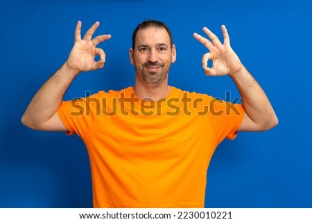 Happy smiling hispanic man with a beard wearing a casual orange t-shirt doing the ok gesture with his two hands celebrating something, isolated on blue studio background.