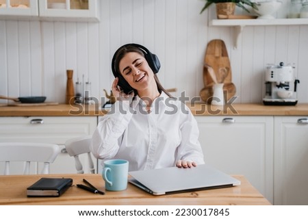 Beautiful brunette young woman in white shirt sitting at table with laptop eyes closed listening music using headphones. Pretty hispanic girl at break of remote learning. Domestic leisure and music