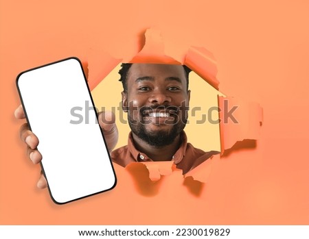 Nice offer. Happy young african american man posing in torn hole of orange paper wall, holding cell phone in hand, showing white blank screen, mockup empty space for advert, collage