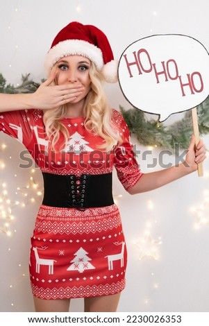 Beautiful blonde woman in a red Christmas dress poses against the background of Christmas tree branches and a glowing garland. Holds a sign in his hand that says Ho-Ho-Ho