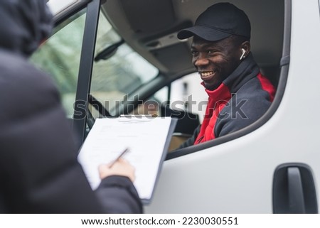 Happy positive Black courier joking around with unrecognizable person taking a delivery. Unrecognizable person signing delivery documents standing next to white delivery truck. High quality photo