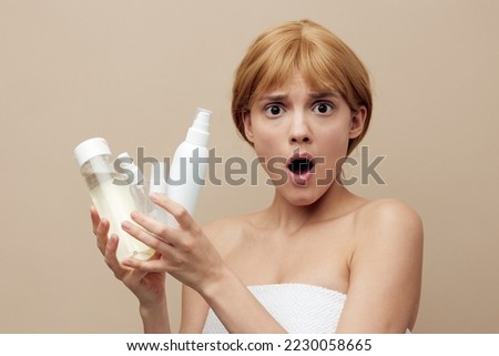 a surprised, shocked woman stands on a beige background holding various jars of care cosmetics in her hands and looks at the camera with her mouth wide open. Horizontal photo with empty space