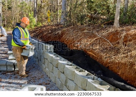 Contractor installing concrete block wall as part of retaining wall construction that is being built as part project Royalty-Free Stock Photo #2230067533