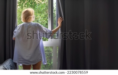 A woman in a man's shirt opens blackout dark curtains Royalty-Free Stock Photo #2230078559