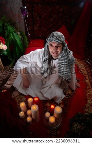 Arab sheikh in a white robe and a headscarf in a boudoir in a room with a red carpet and candles with smoke. Oriental eastern fabulous style photo shoot with a man. Male model posing in fary tale