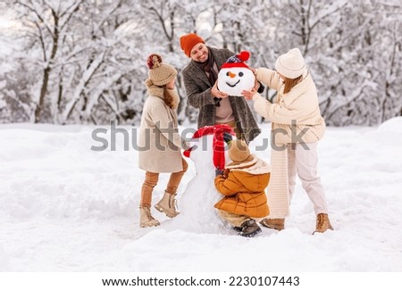 Happy parents and children gathering in snow-covered park together sculpting funny snowman from freshly fallen snow. Father, mother and two kids playing outdoor in winter forest. Family active holiday Royalty-Free Stock Photo #2230107443