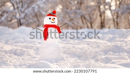 Cheerful snowman in red knitted hat and scarf with carrot nose and joyful smile on his snowy cartoon face standing in winter park against backdrop of snow-covered trees. Fun winter outdoor activities