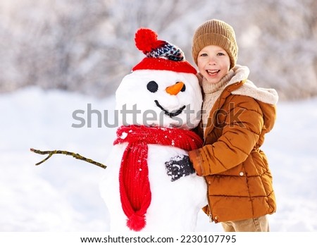Happy child cute little boy in warm clothes with snowman in snowy winter park on beautiful sunny frosty day, selective focus. Snow games for children, outdoor wintertime activities for kids