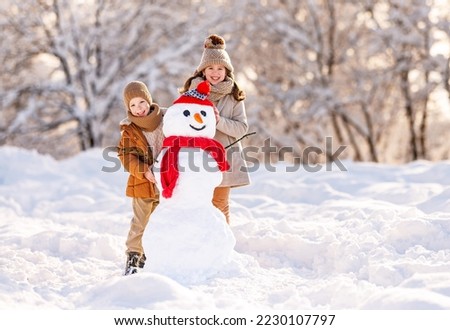 Happy children little brother and sister dressed in warm stylish clothes standing in winter snowy park broadly smiling while proudly posing near snowman made by them, kids playing with snow outdoors