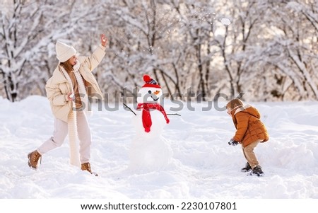 Happy overjoyed family mother and little son play snowballs outdoors in wintertime while standing near snowman, mom and kid laughing, having fun and playing with snow in snowy winter park