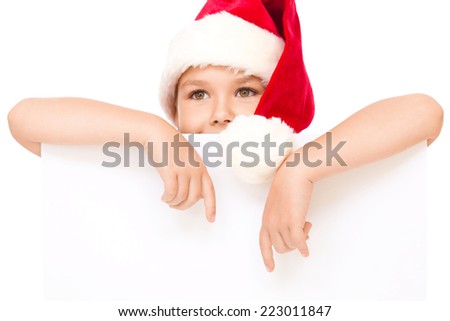 Little girl in santa hat is holding blank board and pointing to it using index finger, isolated over white