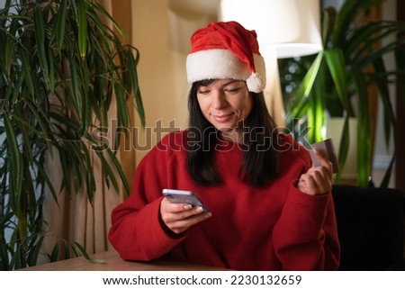 Happy woman holding smartphone and banking credit card, online mobile shopping at home, happy female in Santa hat shopper purchasing goods or services in internet. Copy space