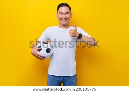 Cheerful young Asian man football fan in a white t-shirt holding a soccer ball and showing thumbs up gesture isolated on yellow background. People sport leisure lifestyle concept