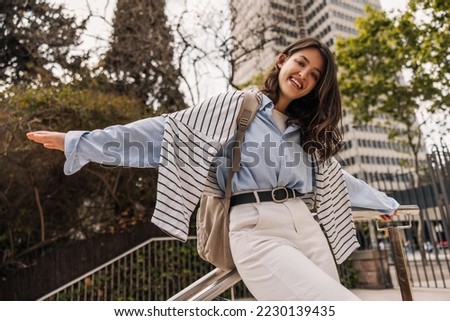 Cheerful young caucasian girl smiling looking at camera, waving her arms walking outdoors. Brunette wears shirt, sweatshirt, trousers and backpack. Weekend enjoyment concept