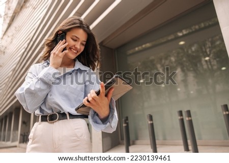 Positive young caucasian woman walking with modern gadgets on street near city business center. Brunette is talking on phone and looking at smartphone screen. Technology concept Royalty-Free Stock Photo #2230139461