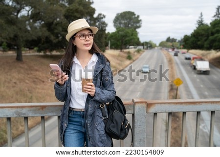 cheerful asian Japanese girl traveler using phone for guide and enjoying cityscape of western America on pedestrian bridge over highway while traveling in palo alto city California usa Royalty-Free Stock Photo #2230154809