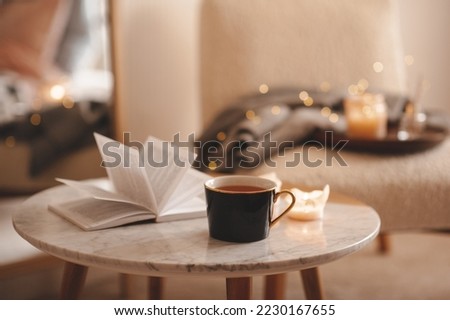 Cup of tea with paper open book and burning scented candles on marble table over cozy chair and glowing lights in bedroom closeup. Winter holiday season. 