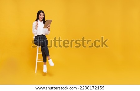 Young woman asian happy smiling. While her using mobile phone sitting on white chair and looking on screen isolate on copy space yellow background. Royalty-Free Stock Photo #2230172155