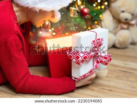 A girl in a red sweater peeks into an open box with a red bow. Christmas photo with a girl in a red cap with a gift from a gift box pours bright light