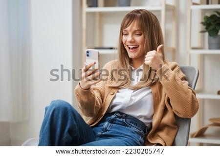 Friendly young blonde lady in warm sweater doing video use phone show thumbs up gesture Like at screen sitting at home. Pause from work, take a break, social media in free time concept. Copy space Royalty-Free Stock Photo #2230205747