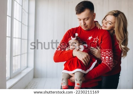Portrait of happy family mom dad and little daughter in red traditional christmas clothes spending time together in light wooden room near window, copy space