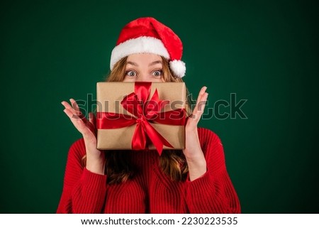 Woman in santa hat covering her face with new year gift isolated on green background. Smiling girl in sweater peeks over christmas box