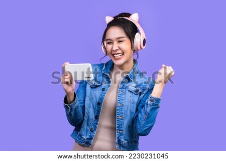 Beautiful Asian young woman in jacket jean and playing video games using joysticks with headphones on voilet background isolated.