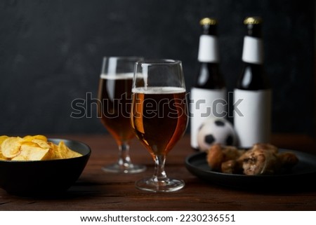 Two glasses with beer, bottles, snacks and soccer ball on wooden table.