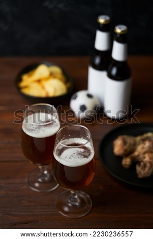 Two glasses with beer, bottles, snacks and soccer ball on wooden table.