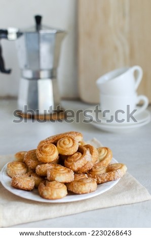 Puff pastry cookies with sugar on a white plate with a geyser coffee maker and two white cups. The concept of homemade baking. Vertical orientation. Selective focus