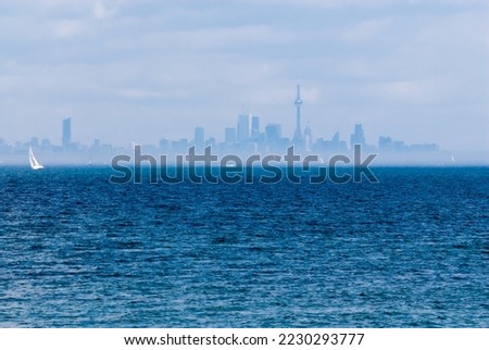 Toronto city skyline from across wavy blue rippled water with sailboat and other boats in distance. City is rising from hazy fog on coastline.