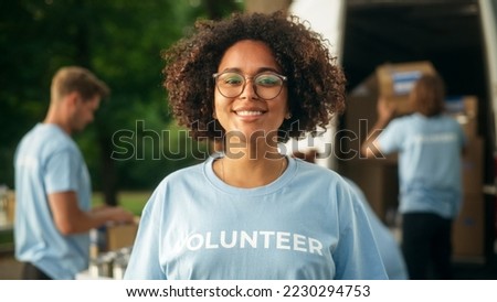 Portrait of a Happy Helpful Black Female Volunteer. Young Adult Multiethnic Latina with Afro Hair, Wearing Glasses, Smiling, Posing for Camera. Humanitarian Aid and Volunteering Concept. Royalty-Free Stock Photo #2230294753